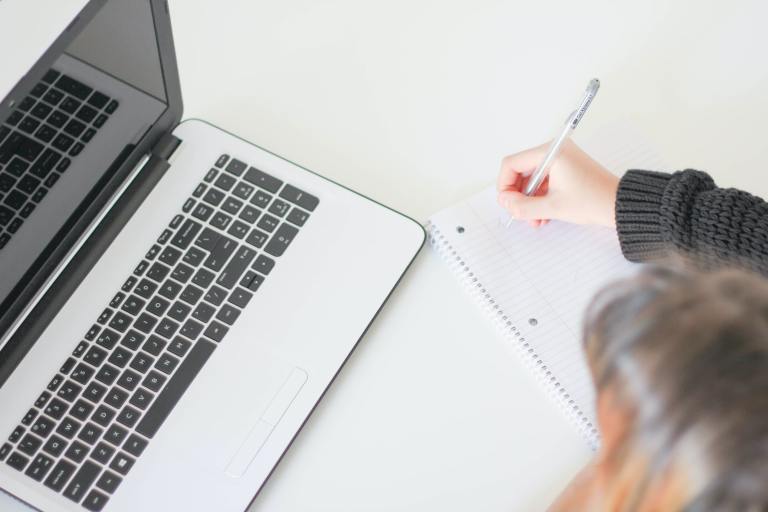 A person writing in a notebook next to a laptop