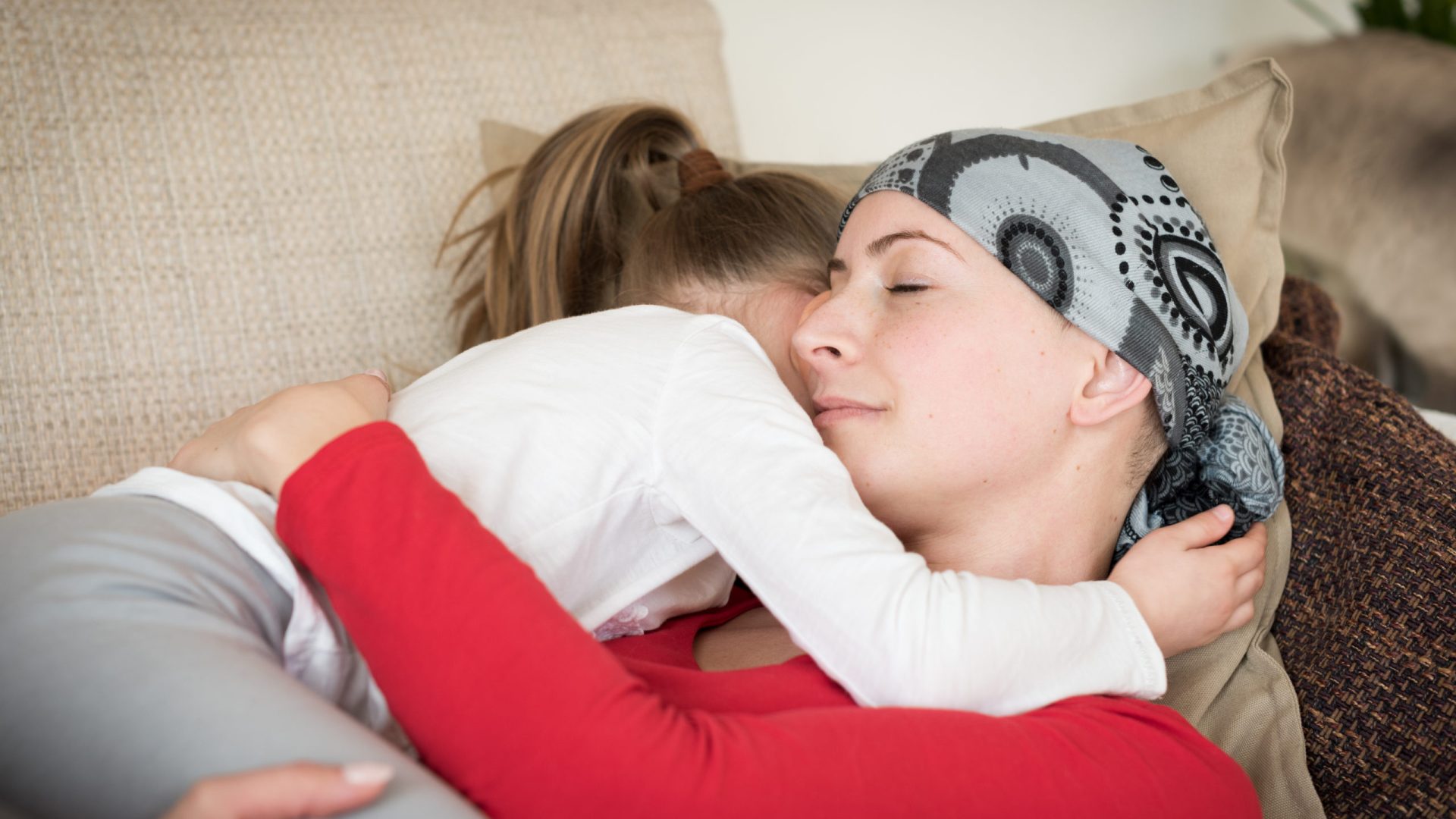 Young adult female cancer patient spending time with her daughter at home, relaxing on the couch. Cancer and family support concept.