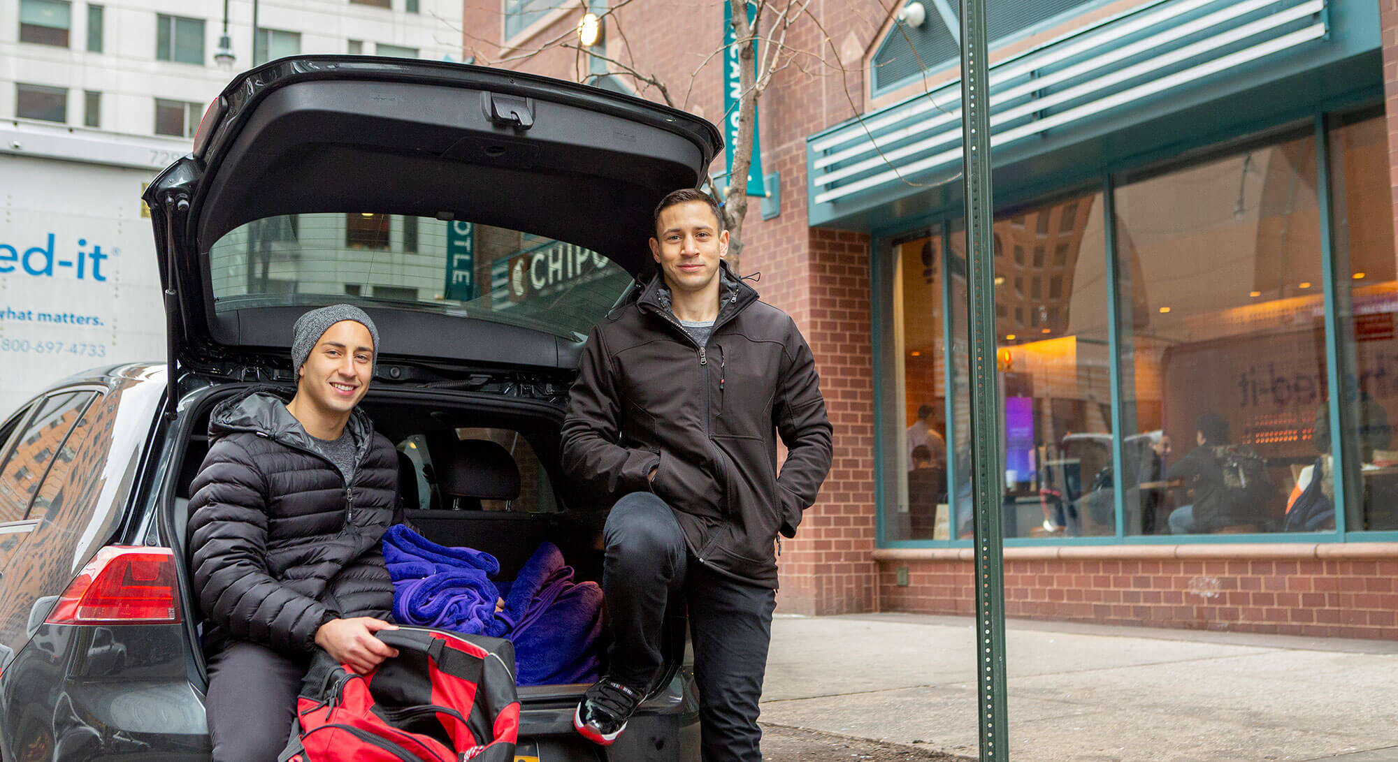 Two man smiling and sitting behind a car