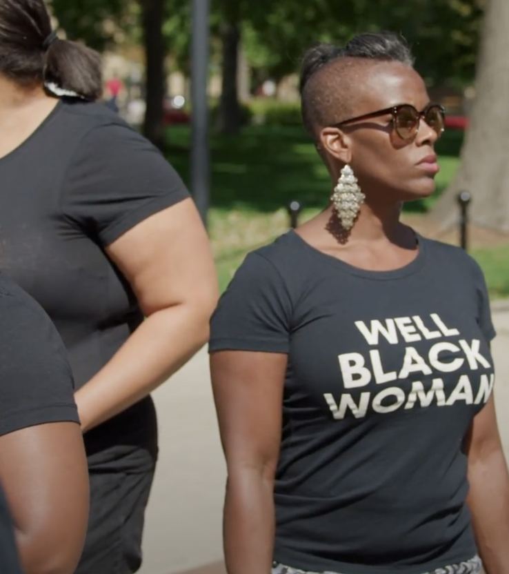 A Black woman in sunglasses and a black t-shirt with white lettering that reads Well Black Woman, outside in the sun, with a group of other Black women