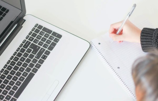 person writing notes in a notebook while using a laptop