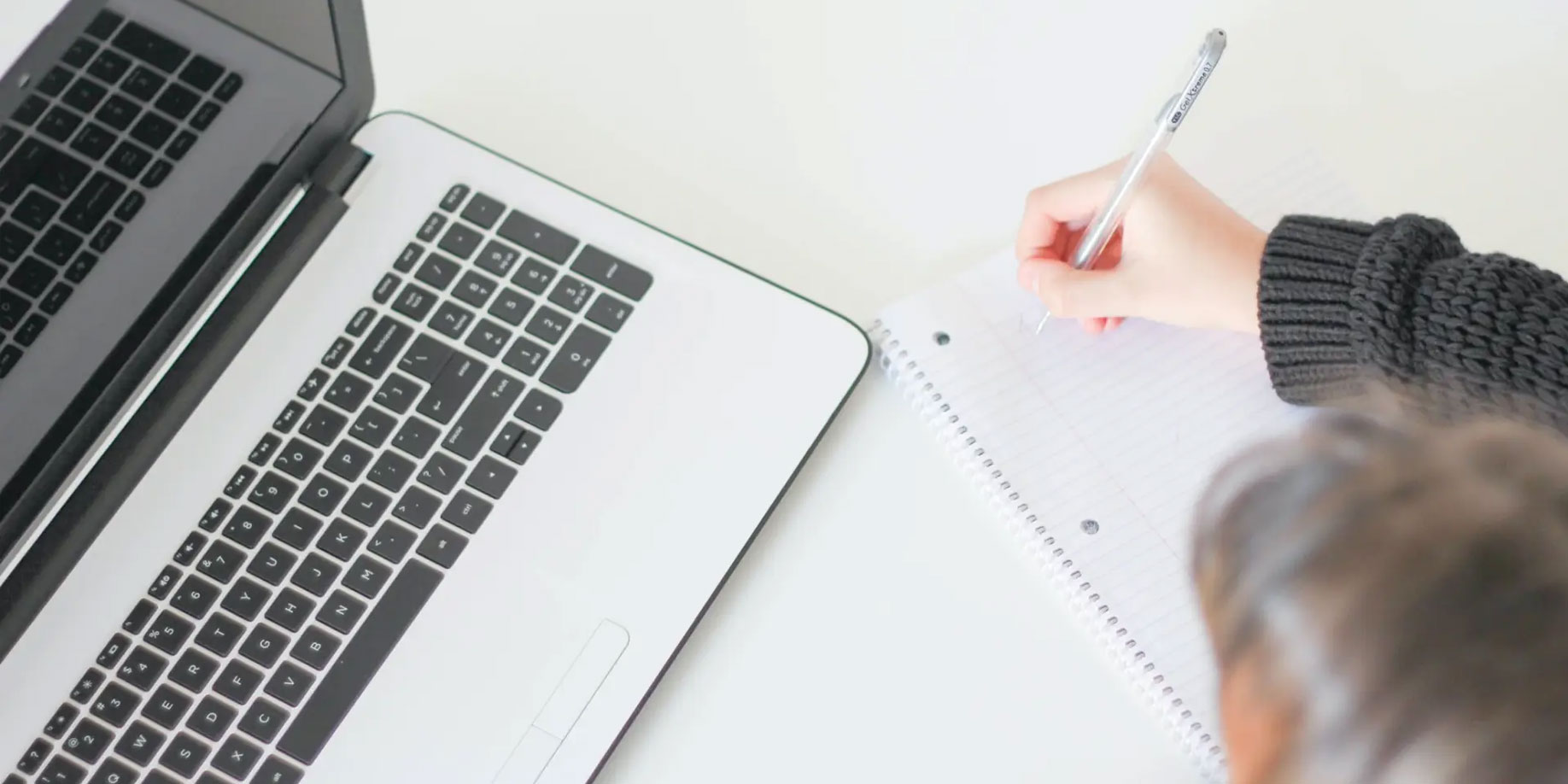 person writing notes in a notebook while using a laptop