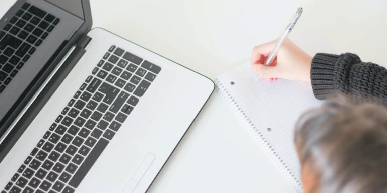 person writing notes in a notebook while using a laptop
