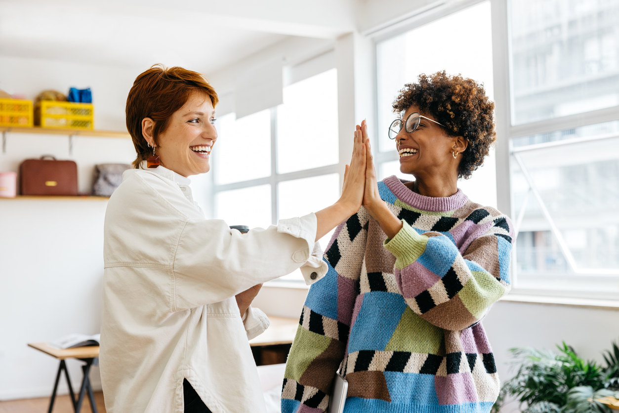 Two happy female nonprofit volunteers high-fiving each other in a bright, modern co-working space, reflecting teamwork and a positive business atmosphere in a freelance office setting.